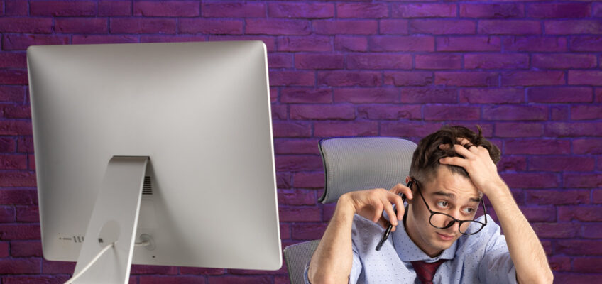 Stressed businessman with glasses looking exasperated while on the phone, sitting in front of a computer screen, symbolizing frustration with the U.S. ESTA website issues.