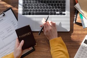 A person filling out an application form next to a laptop, with a passport in hand, preparing for travel documentation such as a U.S. ESTA.