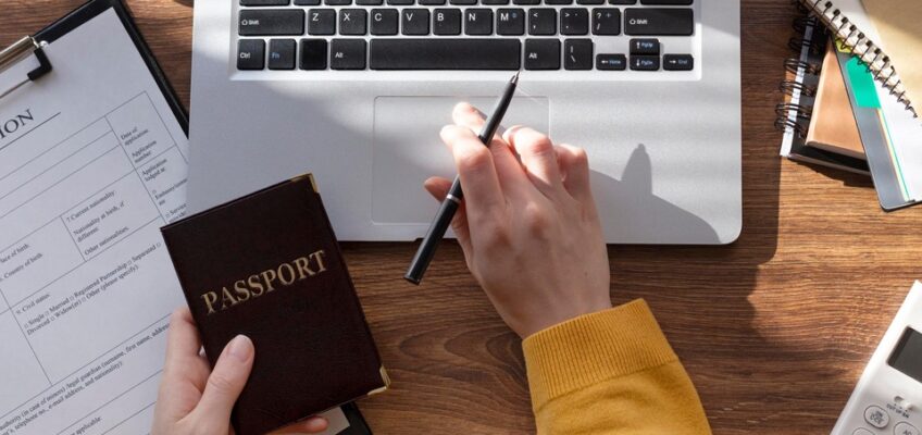 A person filling out an application form next to a laptop, with a passport in hand, preparing for travel documentation such as a U.S. ESTA.