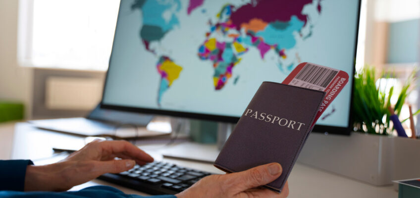 A person holding a passport with an attached boarding pass in front of a computer screen displaying a world map, symbolizing the preparation for international travel with an eVisa.