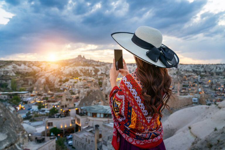 A woman adorned in a vibrant, patterned shawl and a white hat takes a photo with her smartphone, capturing the panoramic view of Cappadocia's unique rock formations and warmly lit caves at sunset.