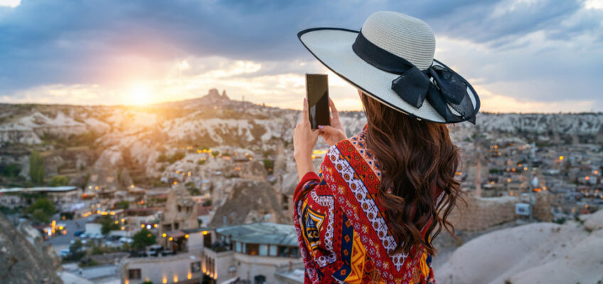 A woman adorned in a vibrant, patterned shawl and a white hat takes a photo with her smartphone, capturing the panoramic view of Cappadocia's unique rock formations and warmly lit caves at sunset.