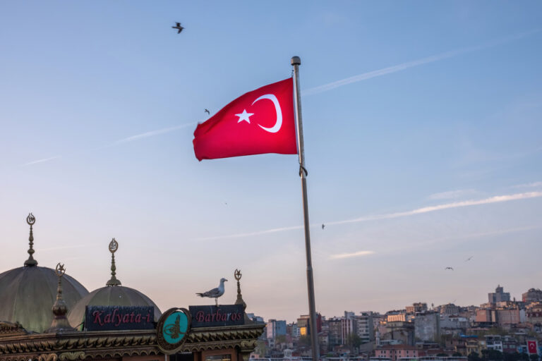 A Turkish flag waving against a sky at dusk, overlooking domed structures and the sprawling cityscape of Istanbul.