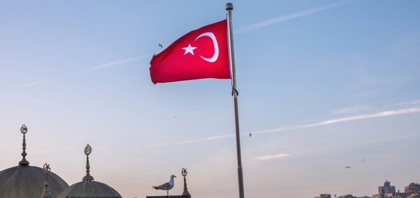 A Turkish flag waving against a sky at dusk, overlooking domed structures and the sprawling cityscape of Istanbul.