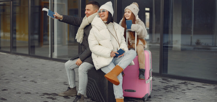 A family of three, smiling and taking a selfie, with a young child sitting on a pink suitcase, all dressed in warm winter clothes, outside an airport, symbolizing group travel.