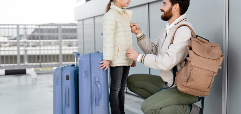 A father, crouched down at an airport, lovingly adjusts his daughter's jacket as they prepare to embark on a journey, with two blue suitcases standing beside them.