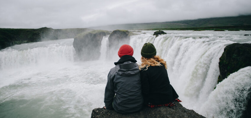 Two travelers sitting on a rock overlooking a powerful waterfall in a misty landscape, embodying the adventure and exploration possible with Canada's eTA multiple-entry travel authorization.