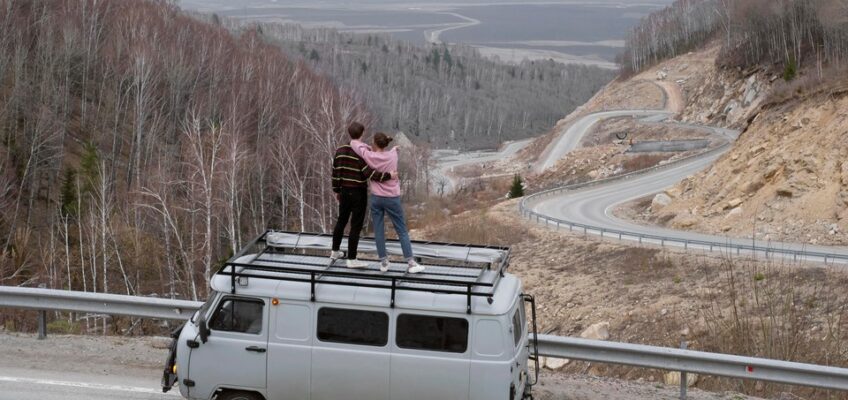 Two people standing on top of a white van parked by the roadside, overlooking a winding road through a leafless forest and hilly terrain.