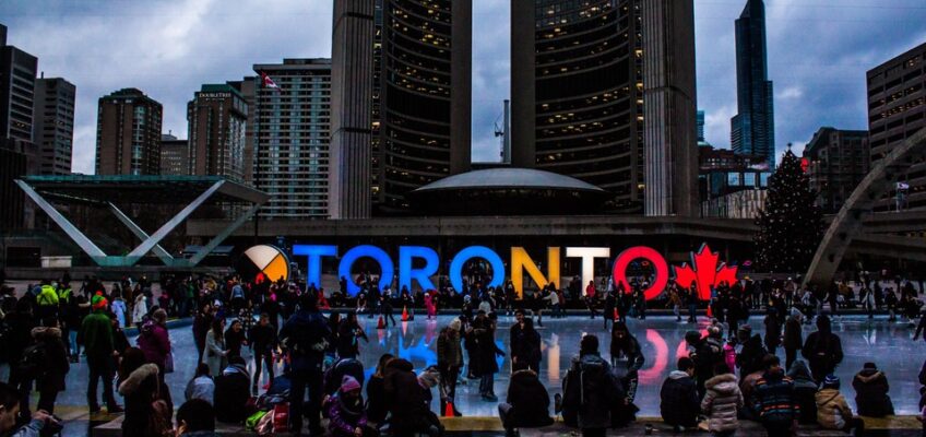 Vibrant scene at Toronto's public square with people ice skating and gathering around the illuminated 'TORONTO' sign with a maple leaf, reflecting the city's dynamic atmosphere.