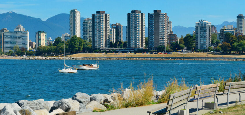 A scenic view of Vancouver's waterfront, with a white sailboat on the water, a line of urban buildings against a backdrop of mountains, and benches along a paved walkway.