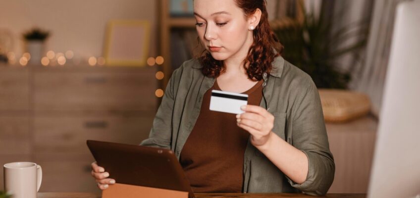 A woman sits at her desk, holding a credit card in one hand and a tablet in the other, attentively making an online payment.