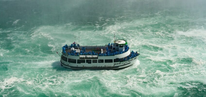 A tour boat, Maid of the Mist, braving the turbulent waters of Niagara Falls, with passengers on board wearing blue raincoats.