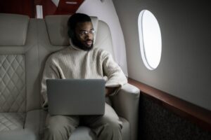 A man in a cream sweater and glasses using a laptop while seated comfortably in the plush leather seat of a private jet, with a window beside him.