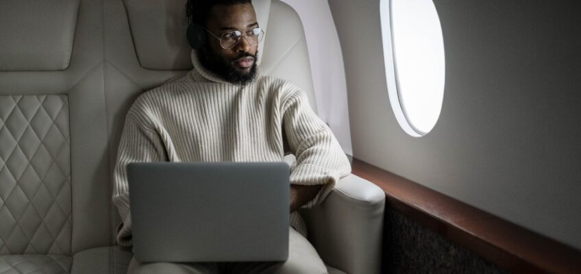 A man in a cream sweater and glasses using a laptop while seated comfortably in the plush leather seat of a private jet, with a window beside him.