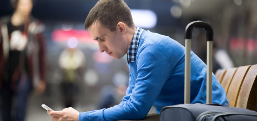 Man in blue sweater sitting with luggage in an airport, anxiously checking his phone.