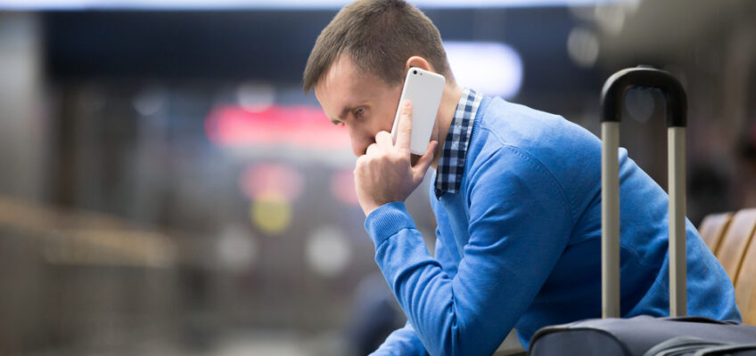 A man sitting in an airport with his suitcase, looking troubled as he talks on his phone, possibly dealing with travel issues.