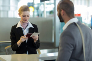 A professional female airport staff member attentively checks a passenger's travel documents at the check-in desk, while a male traveler waits for the verification process to be completed.