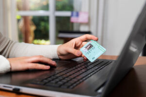 A person holding a green card while using a laptop with an American flag visible in the background, symbolizing the online application process for U.S. travel authorization.