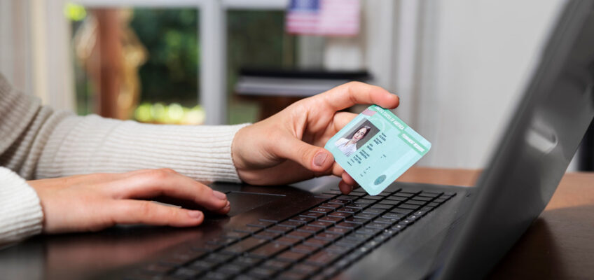 A person holding a green card while using a laptop with an American flag visible in the background, symbolizing the online application process for U.S. travel authorization.