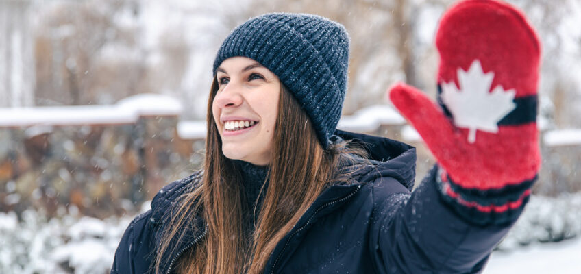 A joyful woman in a winter coat and beanie smiles brightly, waving with a red mitten featuring a white Canadian maple leaf, symbolizing a warm welcome to Canada.