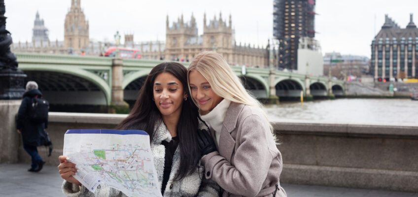 Two women tourists are standing together looking at a map in front of the River Thames in London, with the iconic Houses of Parliament and the under-renovation Big Ben in the background.