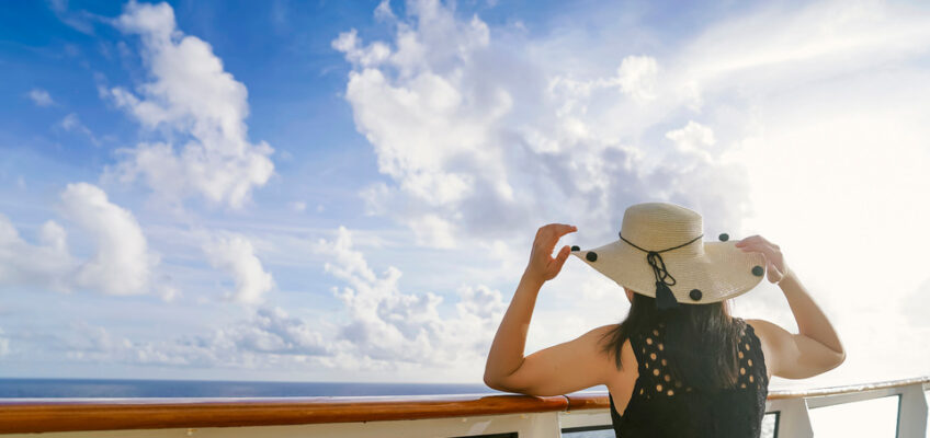 A person holding onto a wide-brimmed hat gazes out at the ocean from the deck of a cruise ship, with a clear blue sky and scattered clouds above.