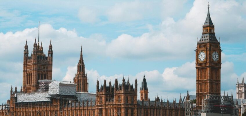 The iconic Houses of Parliament and Big Ben in London, with some scaffolding on one building, as seen across the Thames River with a bridge in the foreground and a red bus passing over.