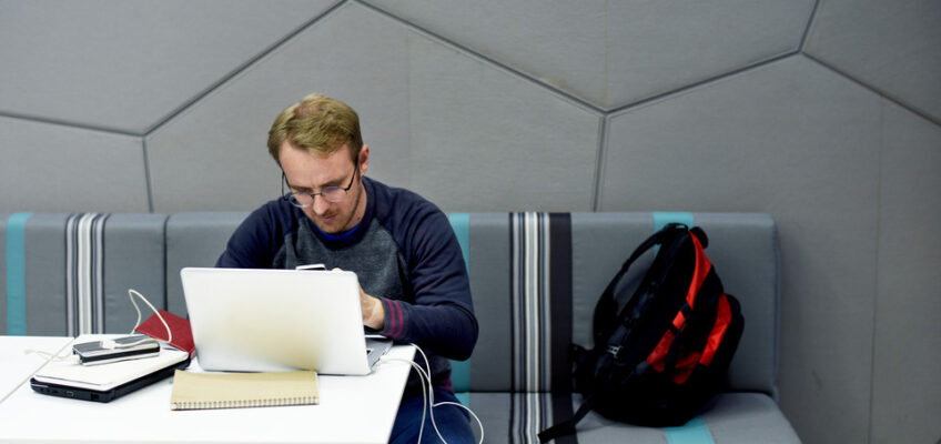 A man intently using a laptop at a modern workstation, with a notepad beside him and a backpack on the adjacent seat, symbolizing diligent travel planning or remote work.