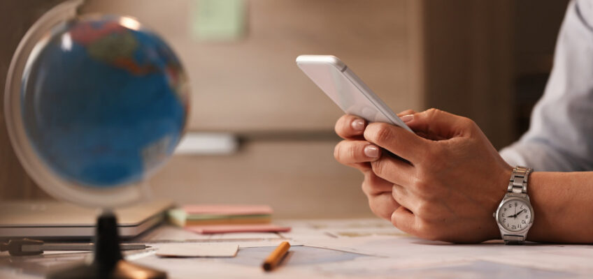 A person sitting at a desk is checking their smartphone, possibly to monitor the status of a UK ETA application, with a world globe, map, and travel planning tools in the foreground, symbolizing trip preparation.