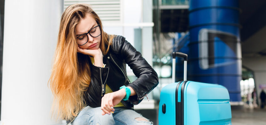 A tired traveler sitting with a suitcase, looking down at her watch, possibly waiting or dealing with a delay.