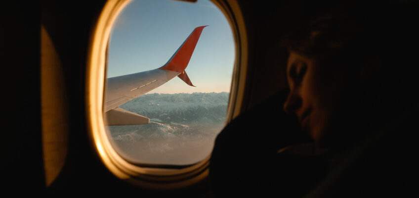 A passenger resting by the window of a plane in flight, with a view of an airplane wing and the glow of sunrise over mountains in the distance.