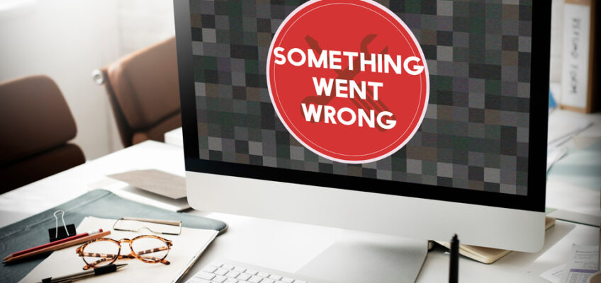 An office desk with an open laptop displaying an error message "SOMETHING WENT WRONG," alongside a notepad, glasses, and writing tools, illustrating technical difficulties.