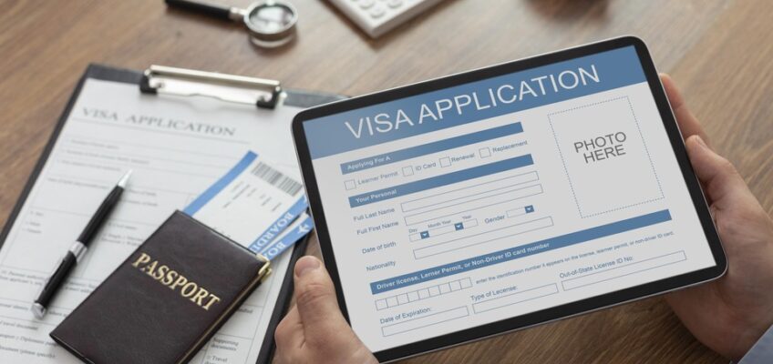 An individual is holding a tablet displaying a visa application screen, with a physical passport, a pen, and a paper visa application form on the desk.