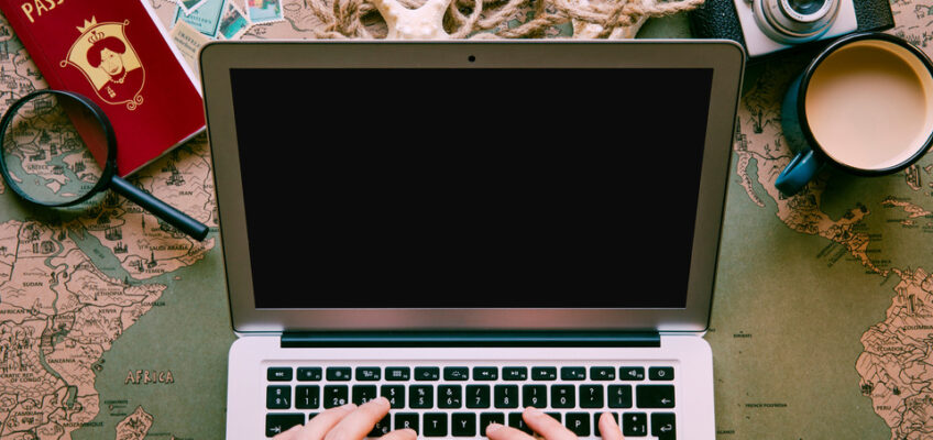 Hands typing on a laptop, surrounded by travel items including a passport, camera, magnifying glass on a world map, and a cup of coffee, representing travel planning and online application completion.