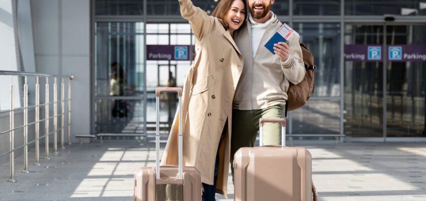 A joyful couple at the airport, with the woman raising her hand in excitement and the man holding up passports, standing next to their luggage, ready to embark on their journey.