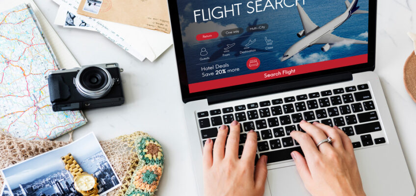 Hands typing on a laptop with a flight search page displayed, surrounded by travel items like a map, camera, and postcards.