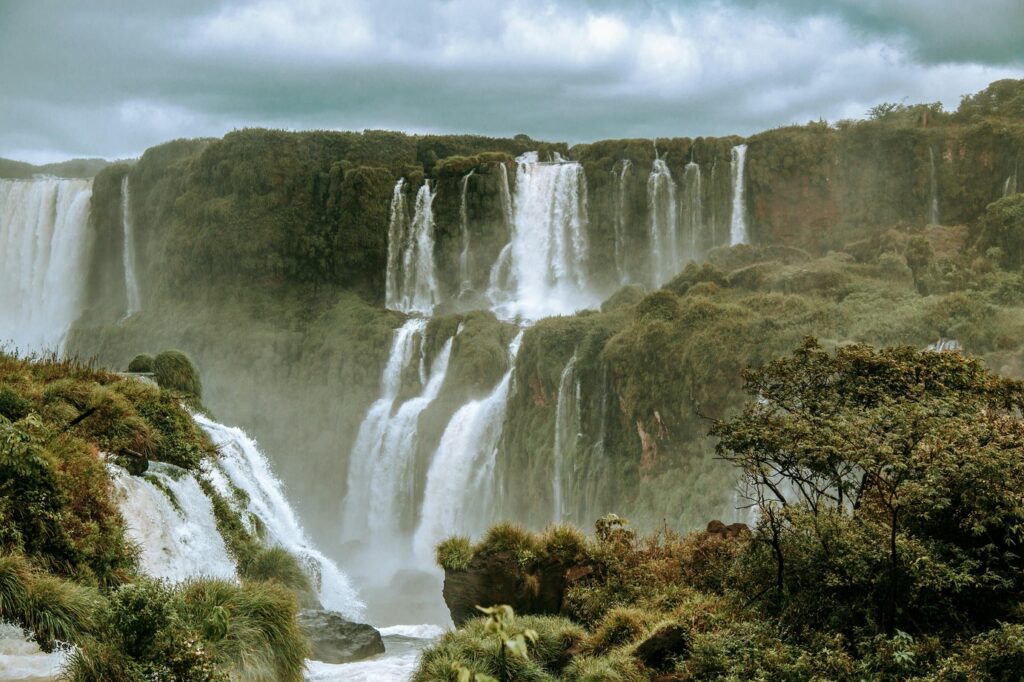 Majestic view of Iguazu Falls with lush green vegetation surrounding cascading waterfalls under a cloudy sky. Mist rises from the powerful flow of water as it tumbles down multiple levels of rocky cliffs, creating a dramatic and vibrant landscape.
