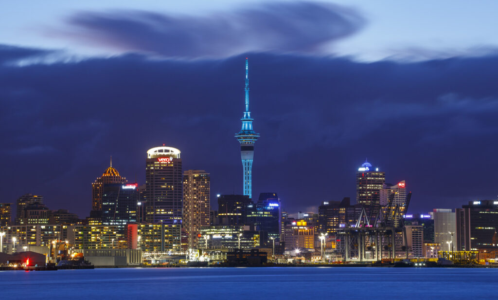 A nighttime skyline view of Auckland, New Zealand, featuring the illuminated Sky Tower and surrounding high-rise buildings, with the waterfront in the foreground.