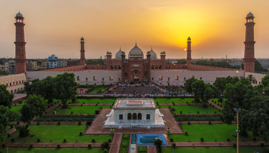 A stunning sunset view of Badshahi Mosque in Lahore, Pakistan, showcasing its grand Mughal architecture with four tall minarets, intricate domes, and expansive courtyard surrounded by lush green gardens and pathways.